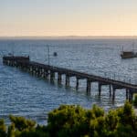 Weathered wooden jetty with railings stretches into deep blue water, two small boats visible beyond, under soft golden light.