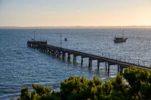 Weathered wooden jetty with railings stretches into deep blue water, two small boats visible beyond, under soft golden light.