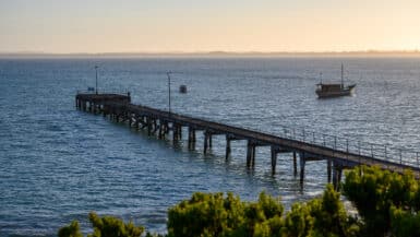 Weathered wooden jetty with railings stretches into deep blue water, two small boats visible beyond, under soft golden light.