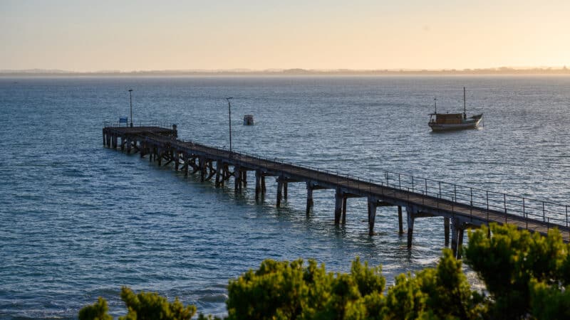 Weathered wooden jetty with railings stretches into deep blue water, two small boats visible beyond, under soft golden light.