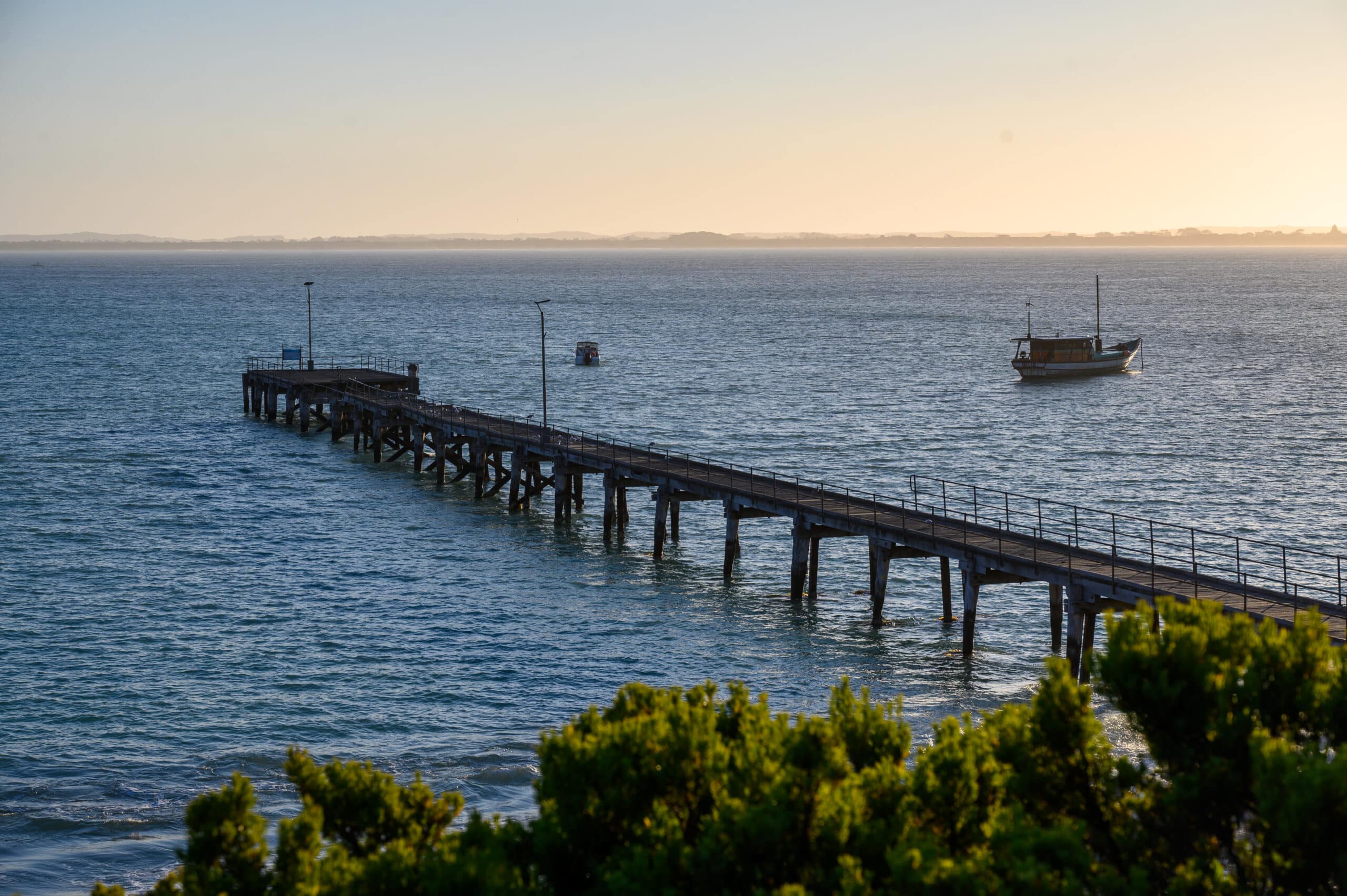Weathered wooden jetty with railings stretches into deep blue water, two small boats visible beyond, under soft golden light.
