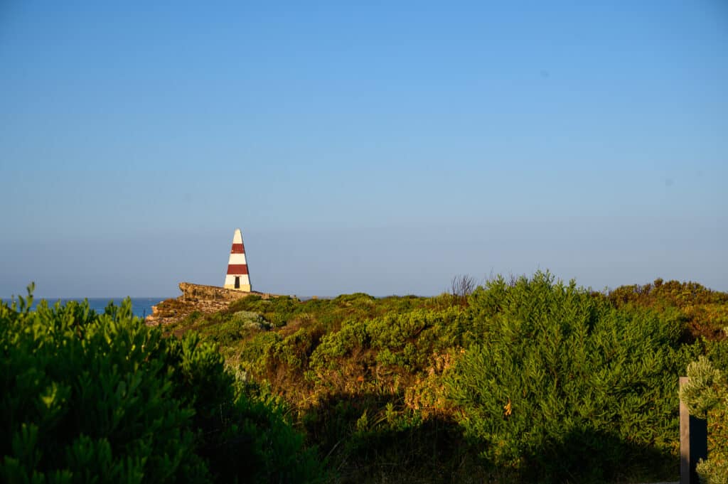 Red-and-white banded Robe Obelisk stands on a rocky headland above coastal scrub under a clear blue sky.