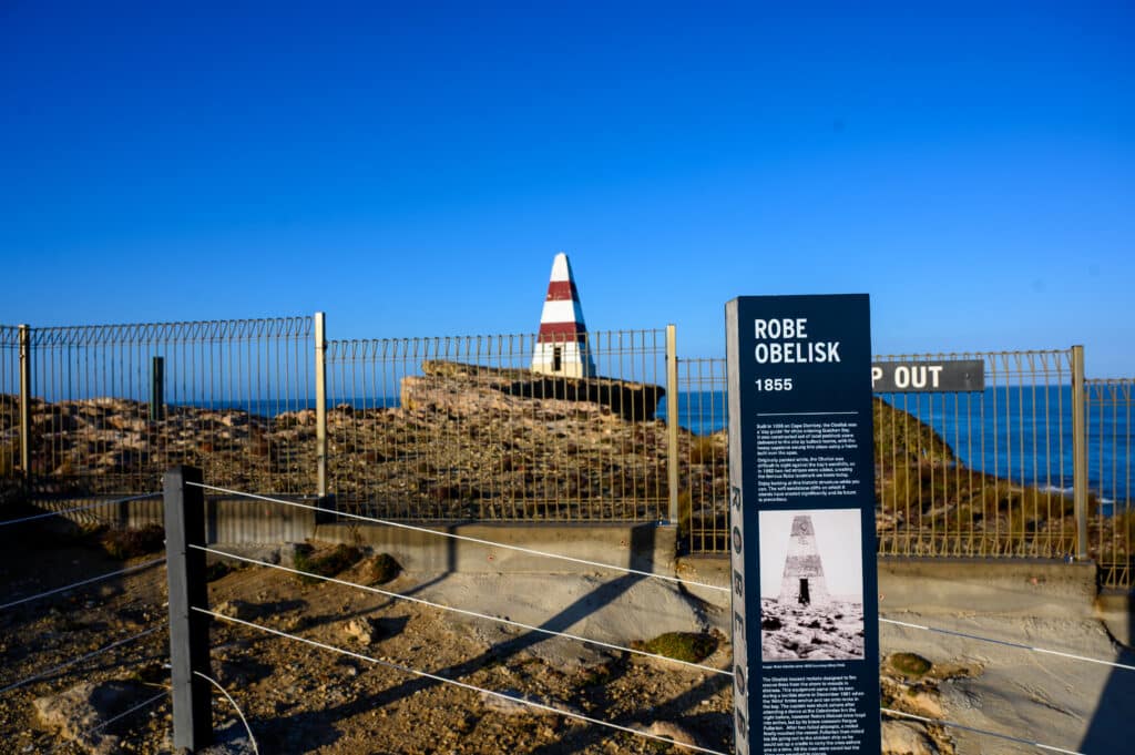 Interpretive sign about the Robe Obelisk stands in front of the red-and-white structure on a fenced rocky headland.