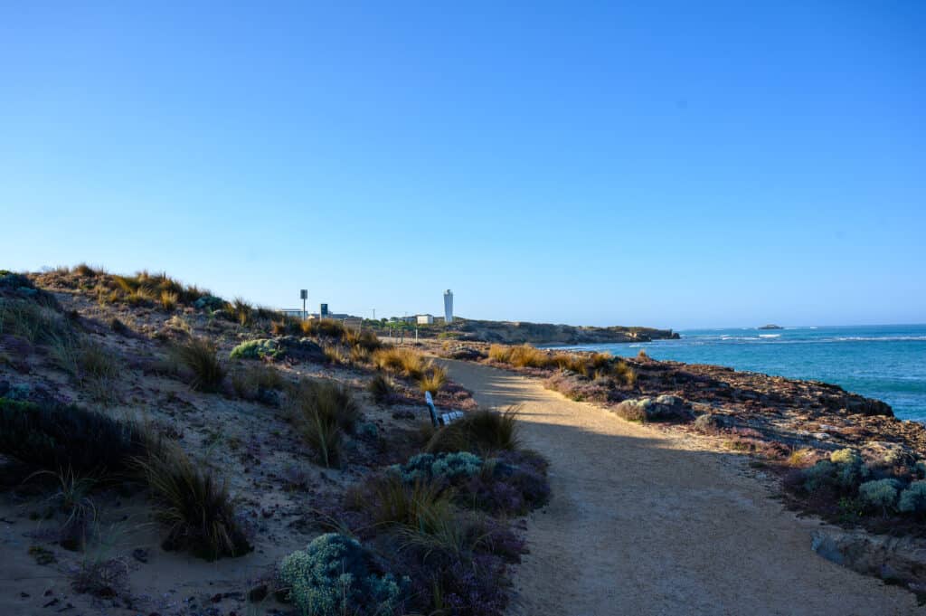 Sandy path winds along a vegetated cliff top toward a distant white tower-like structure, with turquoise sea on the right.