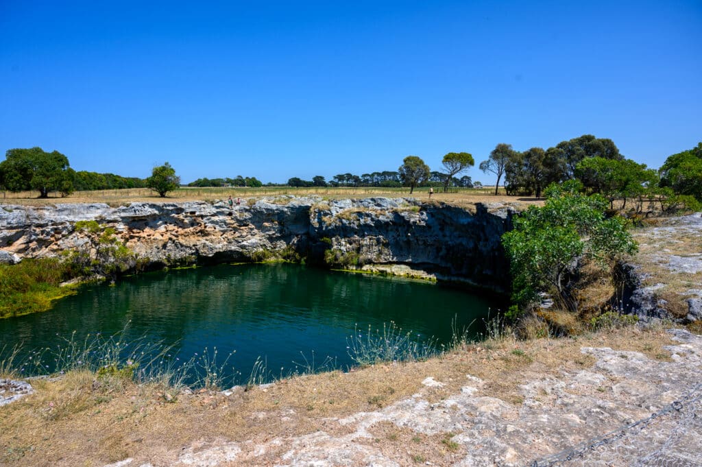 Circular limestone sinkhole filled with deep green water, people standing on the rocky rim under a bright blue sky.