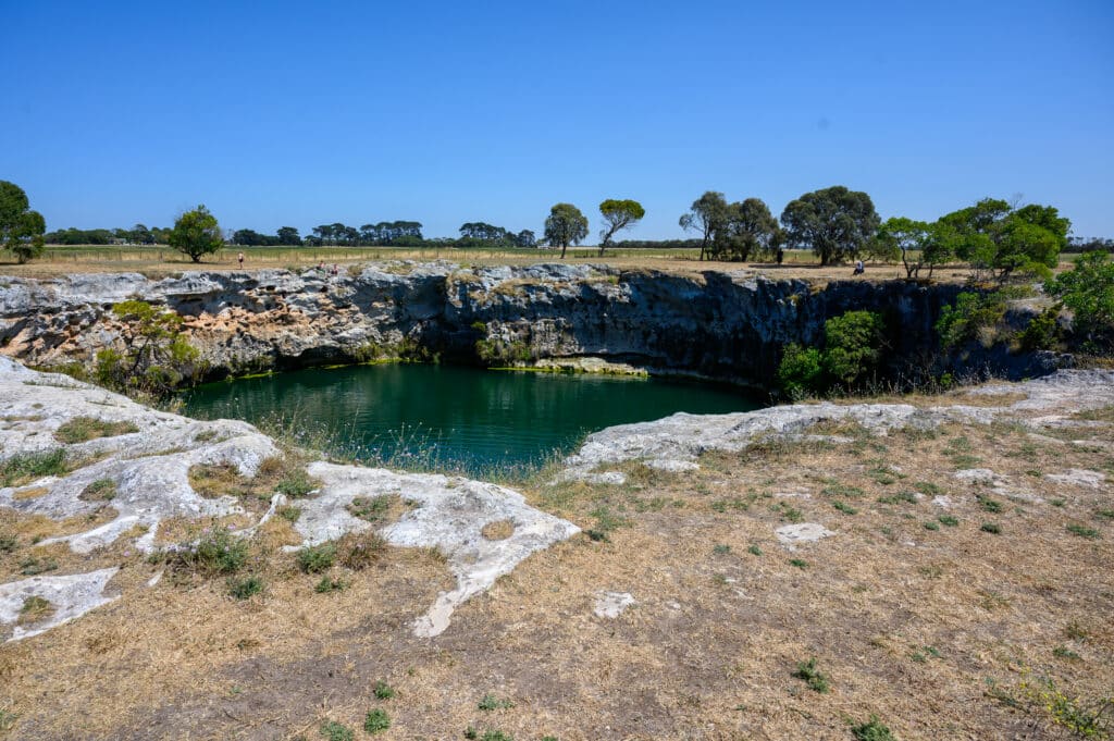Wide view of the green-water sinkhole surrounded by limestone cliffs and dry grassy slopes with visitors on the rim.