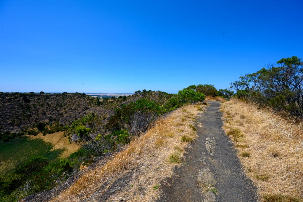 Gravel path runs along the rim of Mount Schank crater, with dry grass and green shrubs overlooking the steep rocky slopes.