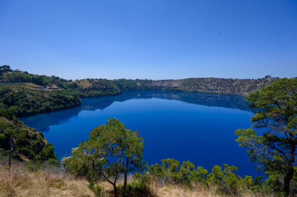 Intense deep-blue water of Blue Lake fills a large volcanic crater, surrounded by tree-covered limestone cliffs under a clear sky.