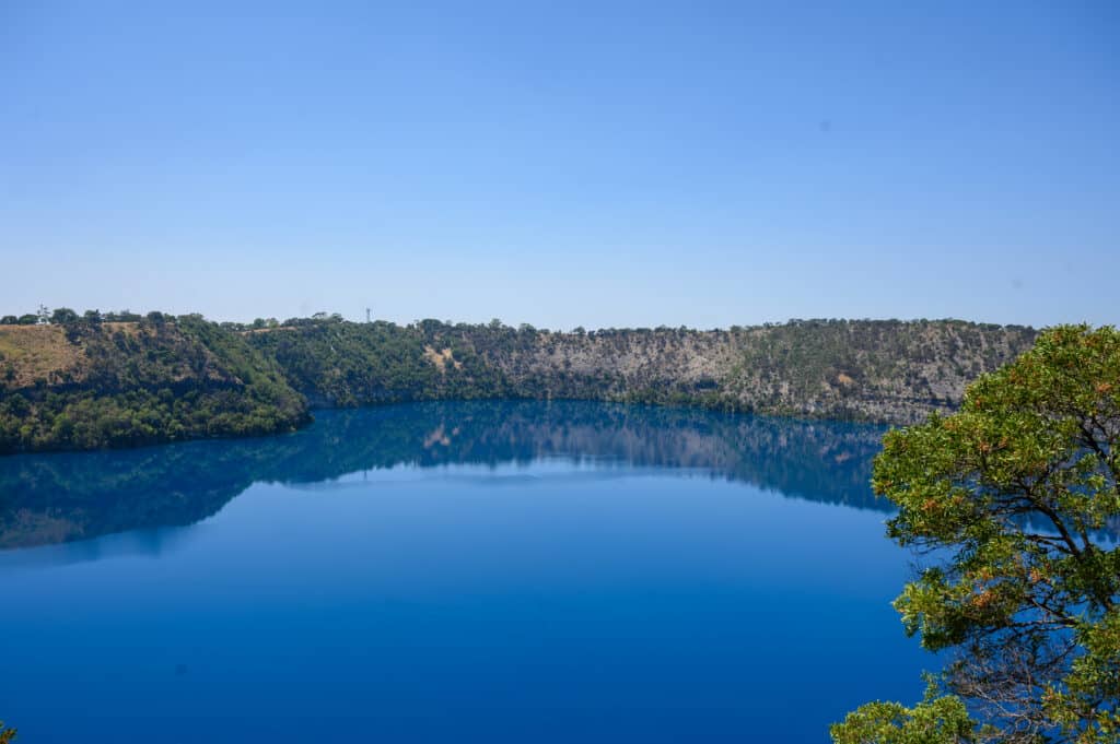 Calm deep-blue surface of Blue Lake reflecting the surrounding tree-lined crater walls under a cloudless sky.