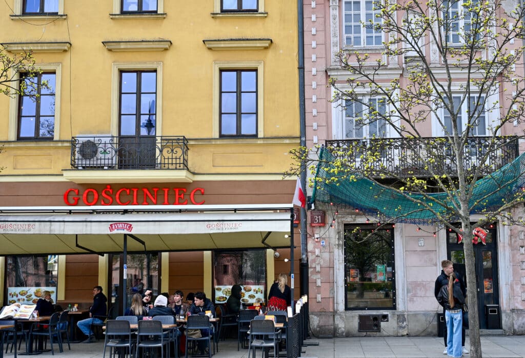 Cafe tables outside a restaurant called Godciniec. Yellow building. Pink stone building to the right