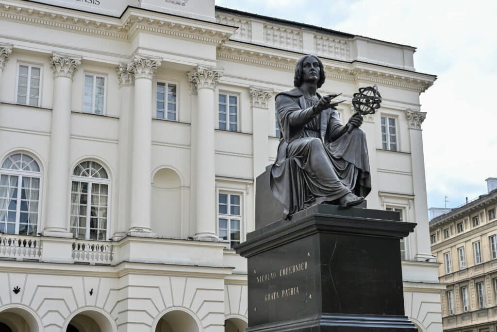 Statue of man holding scientific instruments in front of a white stone building