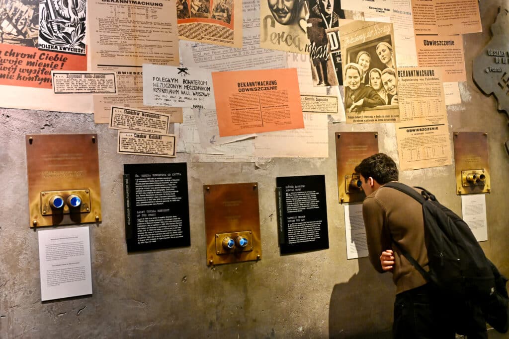 Man peers through binoculars mounted in a wall. Museum display with posters and information pasted on the wall