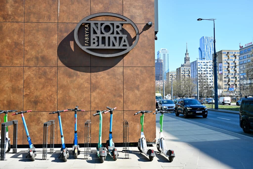 Row of scooters lined up outside Fabryka Norblina building. City buildings to the right