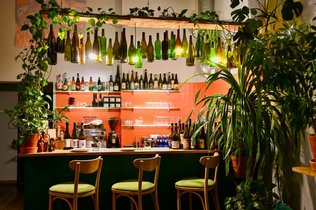 Inside a cafe or restaurant. View of three wooden chairs in front of an orange bar, surrounded by plants and hanging bottles