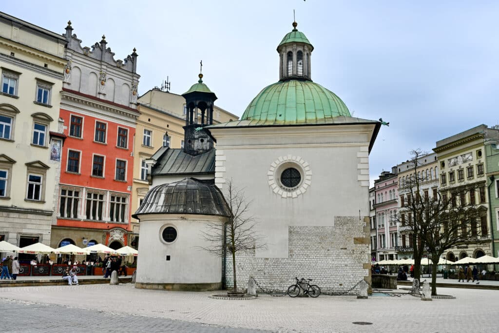 White stone square building with green cupola, circular annex. Colourful townhouses behind.