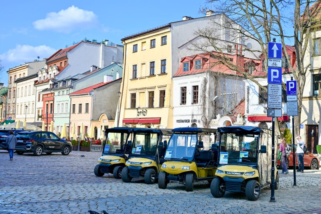 Row of 4 yellow golf buggies parked on cobblestones. Colourful town houses behind.
