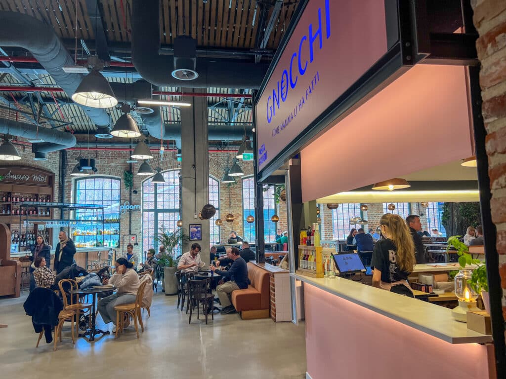 Inside an industrial building with cafes tables inside. Stall advertising Gnocchi to the right