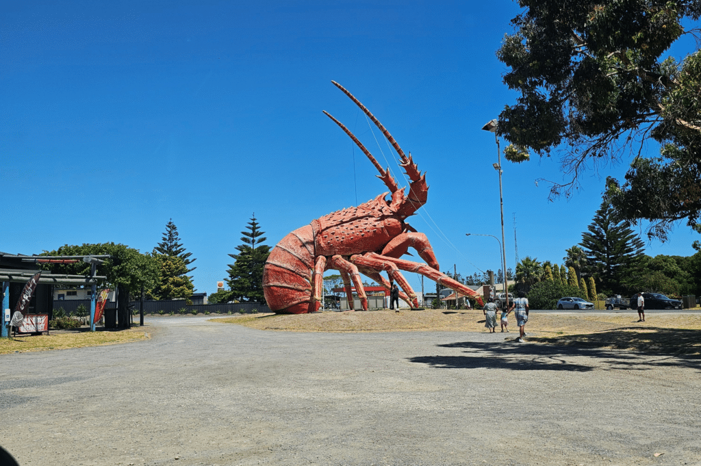 Giant red fibreglass lobster statue known as Larry the Lobster stands on a gravel area in Kingston SE under a clear blue sky, with people walking nearby.
