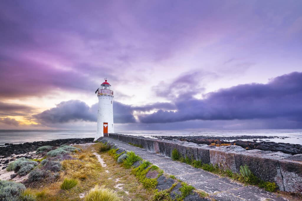 White Port Fairy Lighthouse with red lantern and door stands at the end of a stone path on Griffiths Island, under a dramatic purple and pink cloudy sky at dusk, with rocky shoreline and ocean beyond.