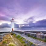 White Port Fairy Lighthouse with red lantern and door stands at the end of a stone path on Griffiths Island, under a dramatic purple and pink cloudy sky at dusk, with rocky shoreline and ocean beyond.