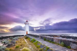 White Port Fairy Lighthouse with red lantern and door stands at the end of a stone path on Griffiths Island, under a dramatic purple and pink cloudy sky at dusk, with rocky shoreline and ocean beyond.