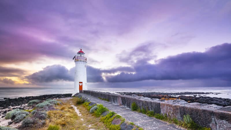 White Port Fairy Lighthouse with red lantern and door stands at the end of a stone path on Griffiths Island, under a dramatic purple and pink cloudy sky at dusk, with rocky shoreline and ocean beyond.