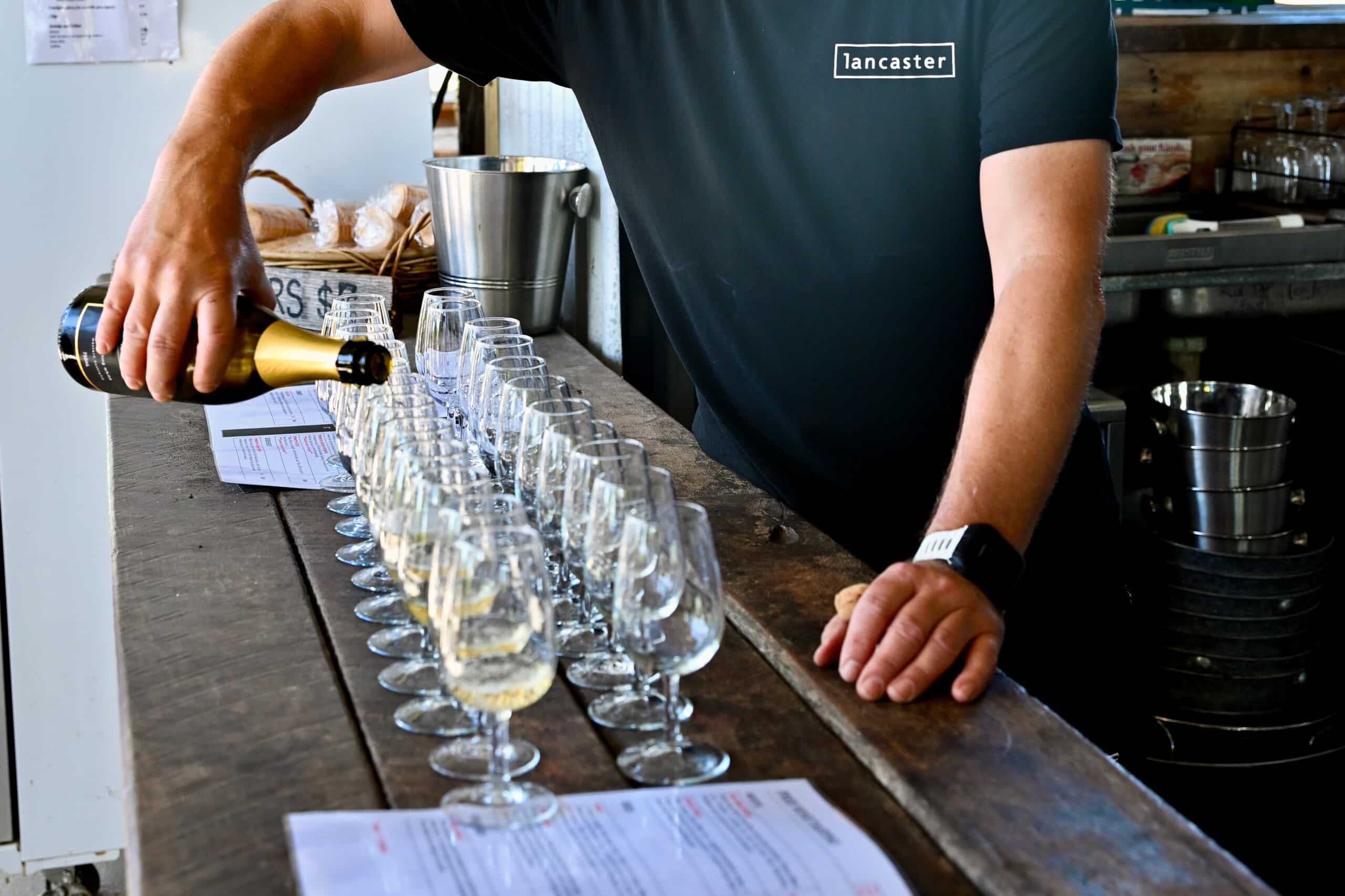 A man in black t-shirt with white logo saying Lancaster is pouring champagne from the bottom into a row of glasses lined up on a wooden bar top. His head is not shown in the photo. Shot at Lancaster Wines, Swan Valley