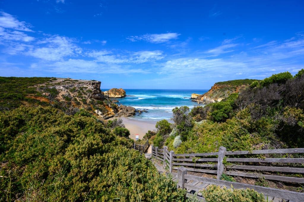 Wooden steps descend through coastal scrub to Childers Cove, with a sandy beach, rugged cliffs and blue ocean in Victoria.