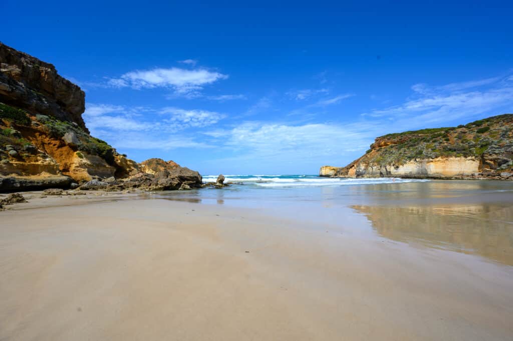 A broad stretch of wet sand between rocky headlands at Childers Cove, with white surf beneath a bright blue sky.