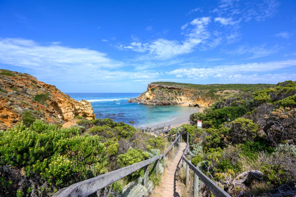 Timber steps lead through green coastal vegetation to Childers Cove, where turquoise water meets a beach beneath layered cliffs.