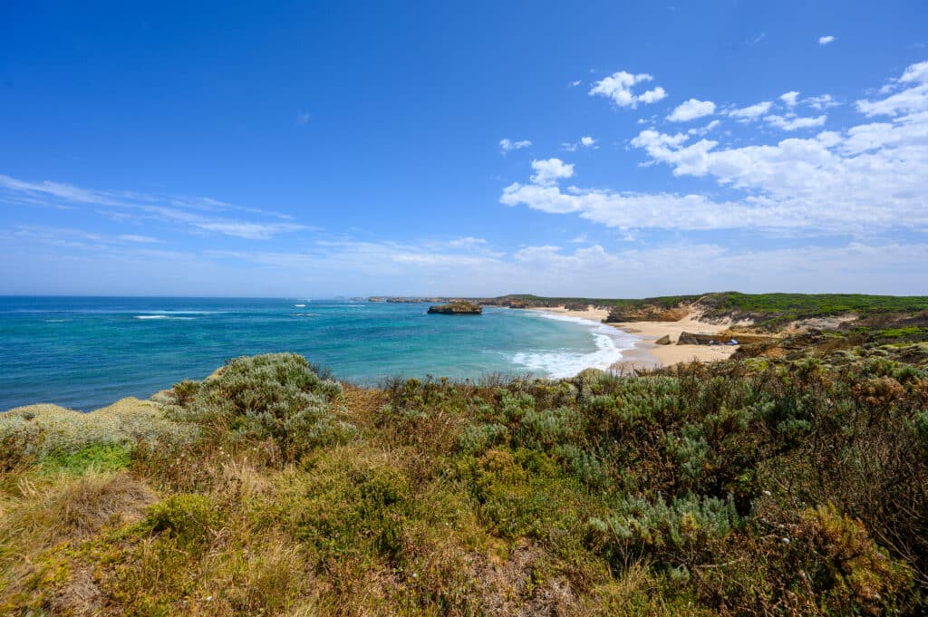 Bay of Martyrs beach curves beneath low cliffs and dunes, with turquoise water, shoreline boulders and an offshore rock.