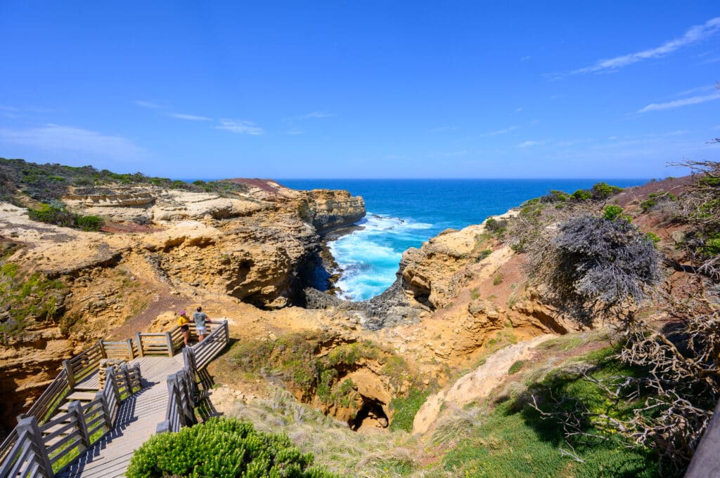 Wooden steps descend between weathered limestone cliffs at The Grotto, with visitors overlooking turquoise surf on Victoria’s coast.