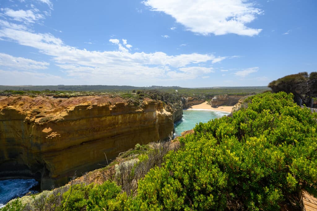The sandy beach inside Loch Ard Gorge lies beyond a narrow turquoise inlet, framed by high limestone cliffs and green scrub.