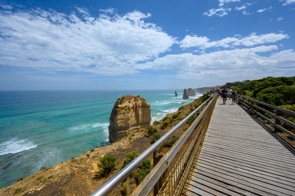 A timber boardwalk follows the cliff edge past a limestone stack towards visitors at the Twelve Apostles lookout.