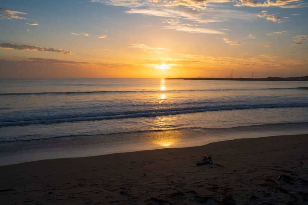 The rising sun casts a golden reflection across waves at Apollo Bay Beach, with harbour masts on the horizon.