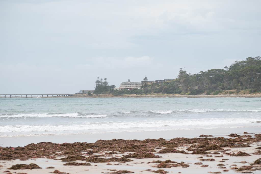 Lorne Pier extends into the bay beside the Grand Pacific Hotel, with breaking waves and seaweed-covered rocks in front.