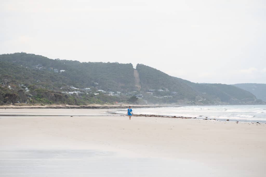 A lone beachgoer in blue walks across Lorne Beach beneath wooded hills dotted with houses.