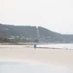 A lone beachgoer in blue walks across Lorne Beach beneath wooded hills dotted with houses.