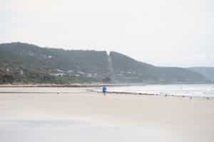 A lone beachgoer in blue walks across Lorne Beach beneath wooded hills dotted with houses.