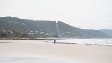 A lone beachgoer in blue walks across Lorne Beach beneath wooded hills dotted with houses.