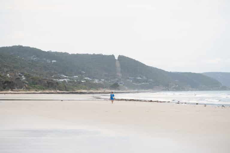 A lone beachgoer in blue walks across Lorne Beach beneath wooded hills dotted with houses.