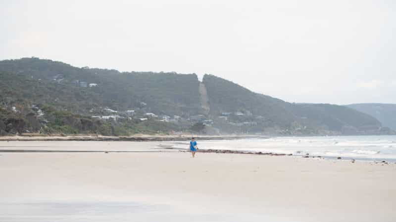 A lone beachgoer in blue walks across Lorne Beach beneath wooded hills dotted with houses.