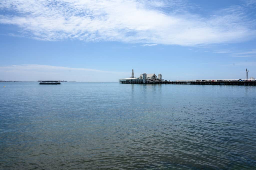 Cunningham Pier stretches across calm Corio Bay in Geelong, with white waterfront buildings beneath a blue, cloud-streaked sky.