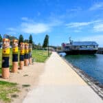 Painted swimmer bollards line Geelong’s waterfront path beside the Geelong Boat House and the blue water of Corio Bay.