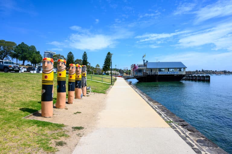 Painted swimmer bollards line Geelong’s waterfront path beside the Geelong Boat House and the blue water of Corio Bay.