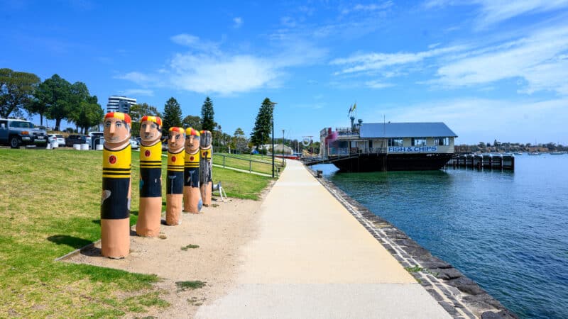 Painted swimmer bollards line Geelong’s waterfront path beside the Geelong Boat House and the blue water of Corio Bay.