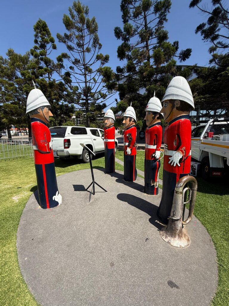 Volunteer Rifle Band bollards in red uniforms and white helmets gather around a music stand on Geelong’s waterfront.
