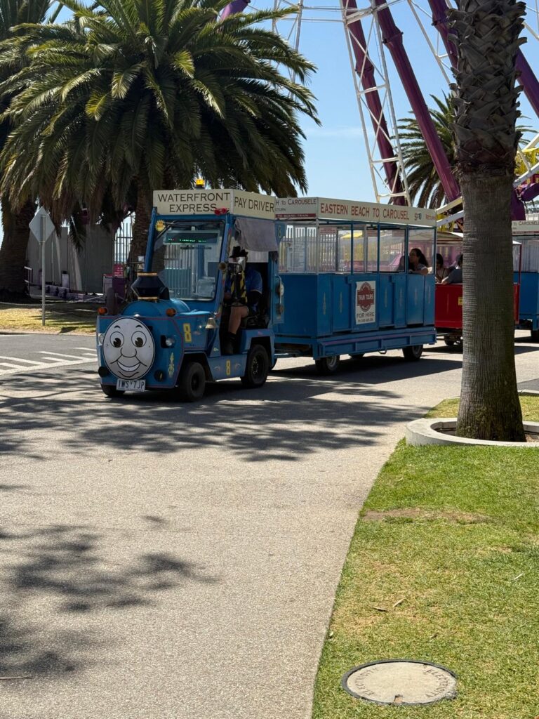 The blue Waterfront Express tourist train sits beside palm trees and the Giant Sky Wheel on Geelong’s waterfront.