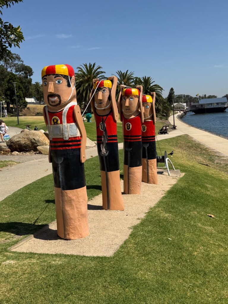 Four Victoria Baths Swimmers bollards wear red-and-black costumes and red-and-yellow caps beside Geelong’s waterfront.