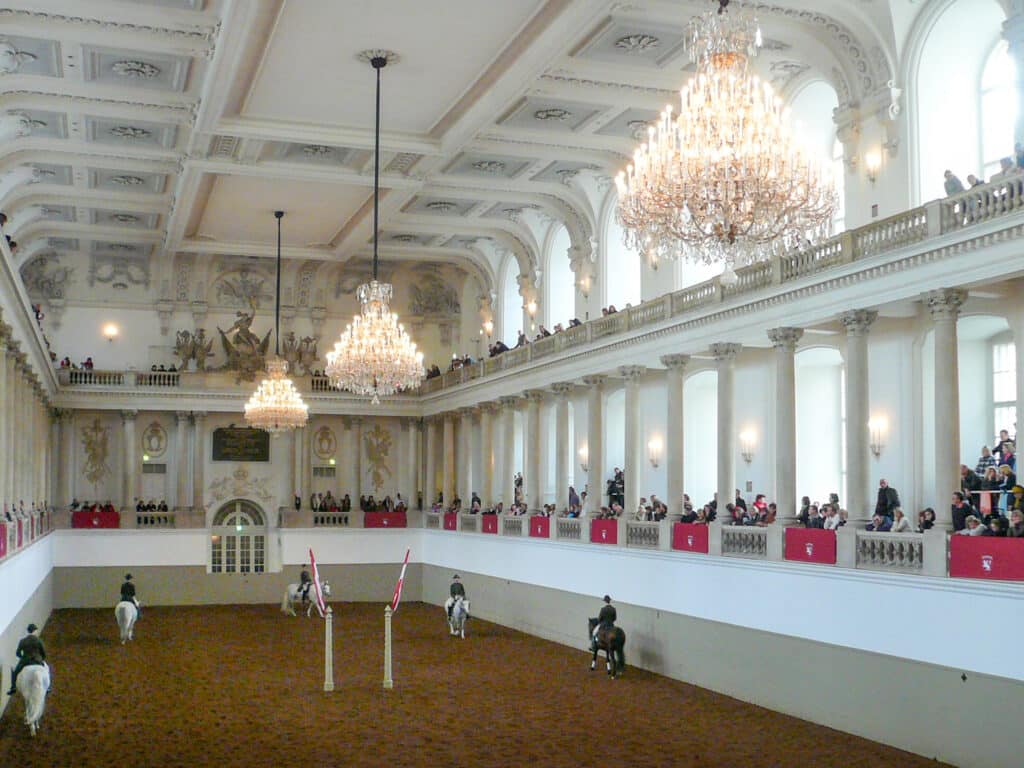 Inside an ornate hall with two tier balconies and chandeliers. Horses walk around on the central floor.